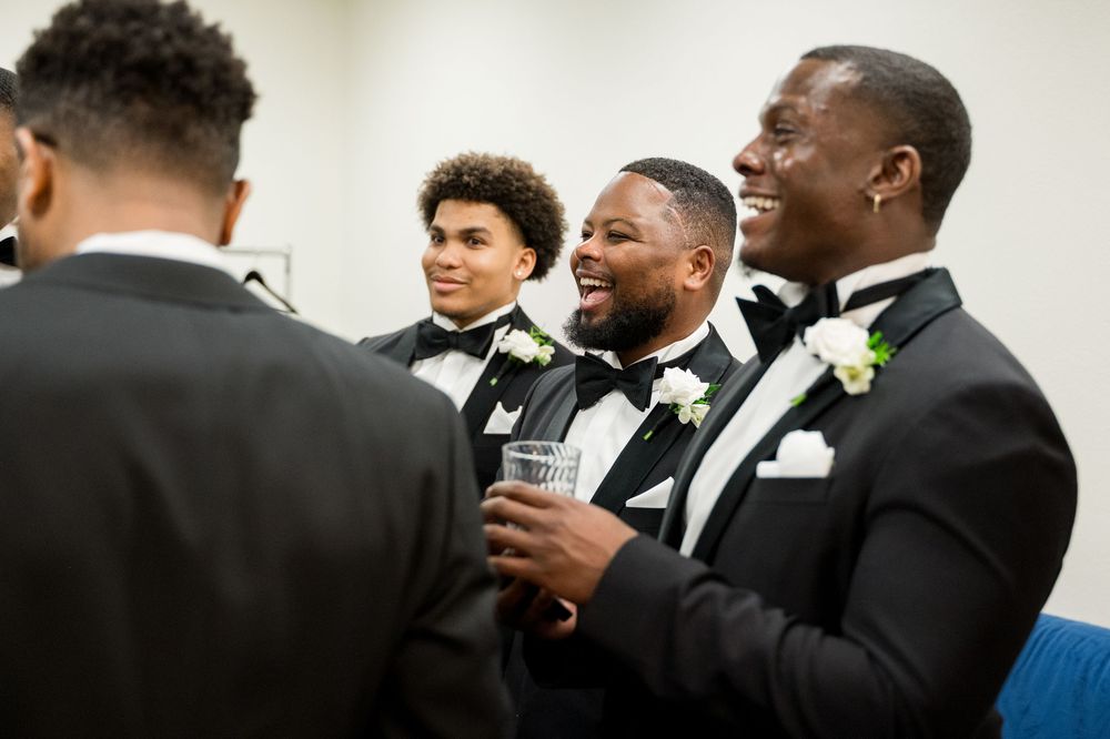 Groomsmen in black tuxedos share a candid laughing moment during a wedding celebration.