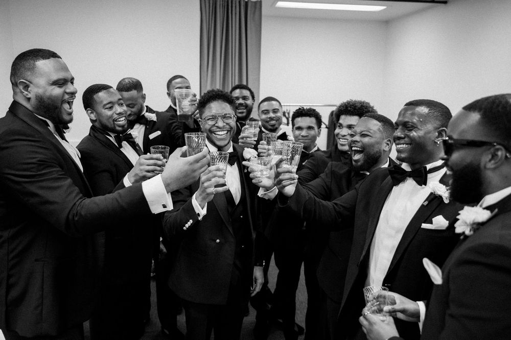 A festive black and white photo showing a group in tuxedos laughing and toasting together in a room.