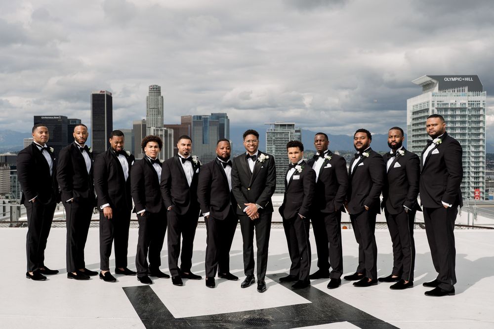 A line of groomsmen in black tuxedos stand on a rooftop against a cloudy city skyline.