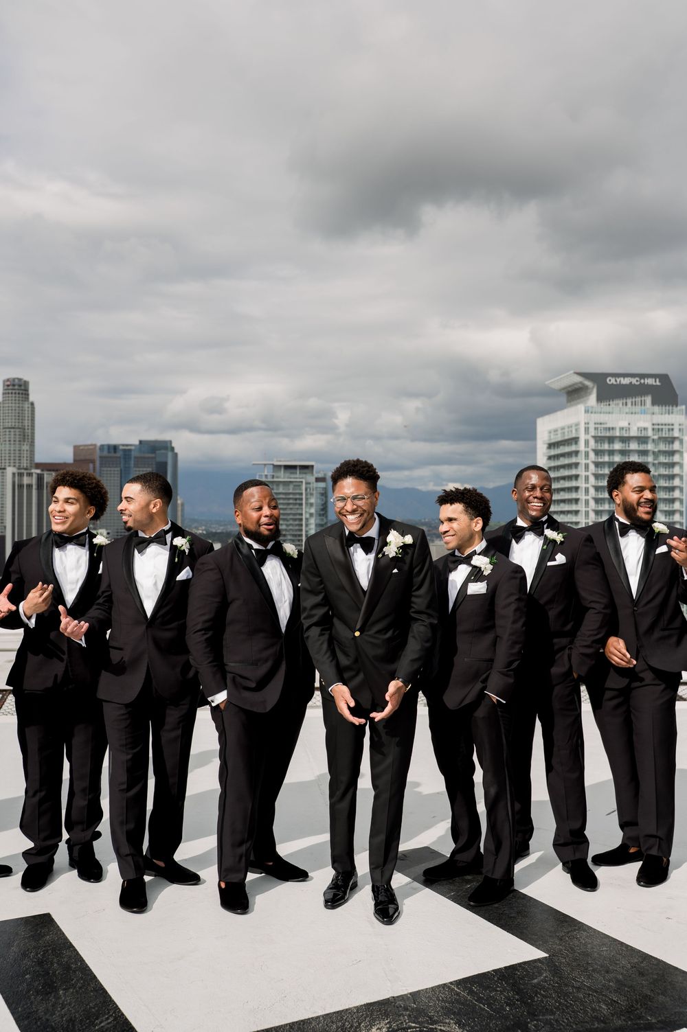 Group of groomsmen in black tuxedos and bow ties posing together on rooftop with urban skyline behind them.