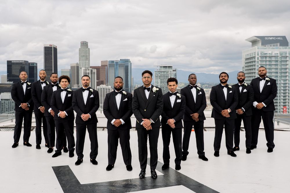 A wedding party in black tuxedos stands on a rooftop with a modern city skyline in the background.