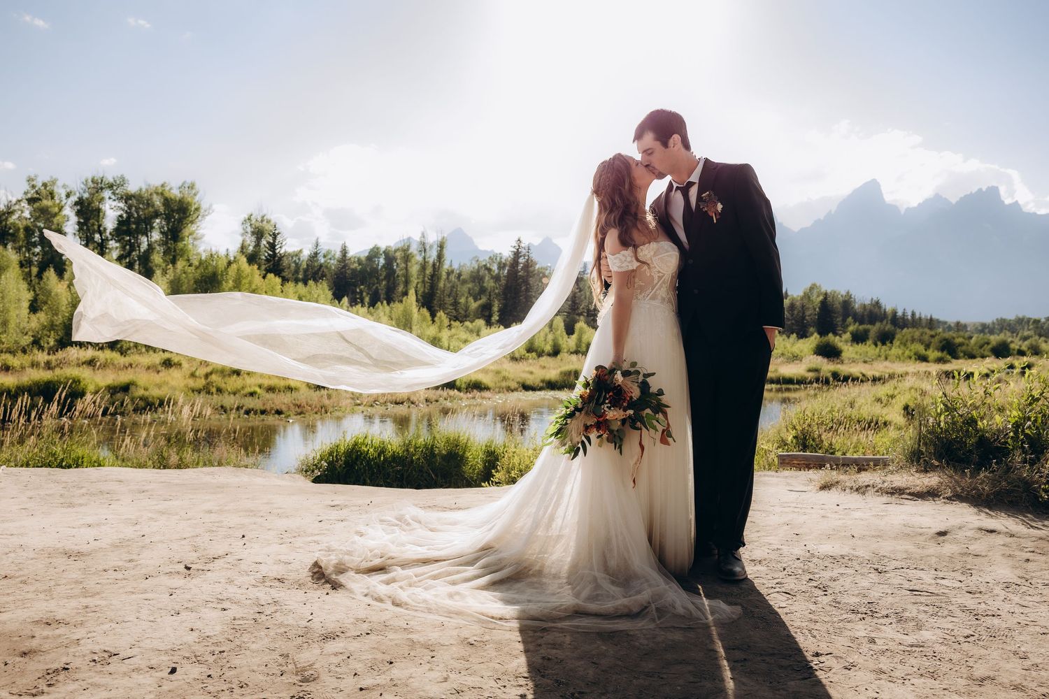 Bride and groom share a joyful moment in golden sunlit field with mountains in background during outdoor wedding ceremony.
