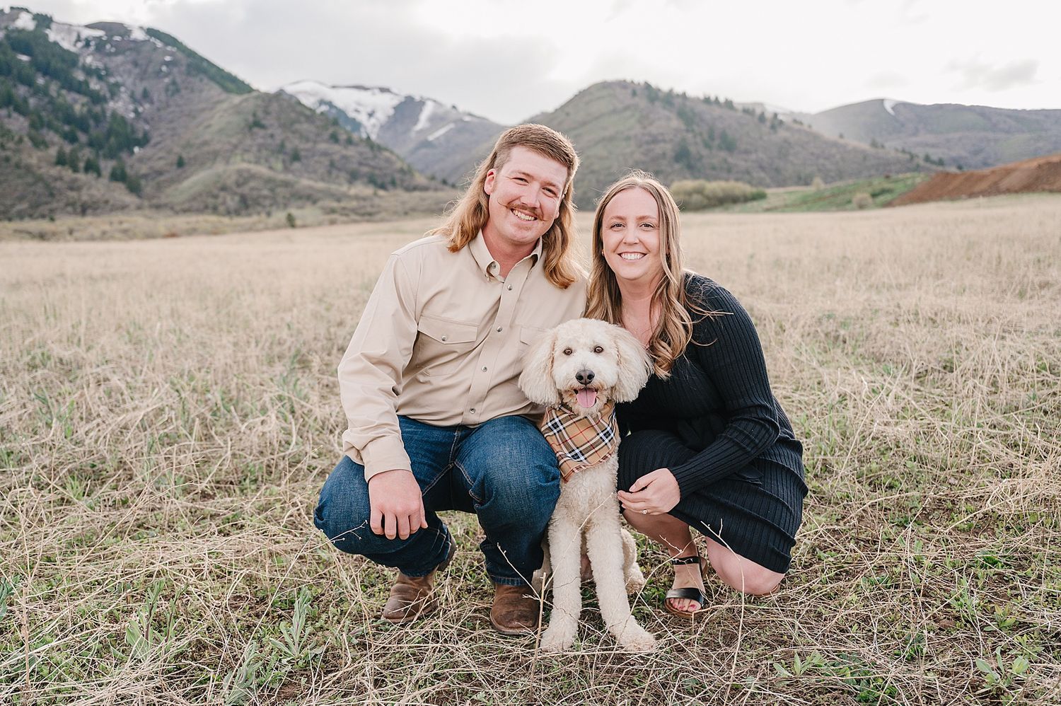 A couple crouches in a grassy field with their white dog as snow-capped mountains rise in the background.