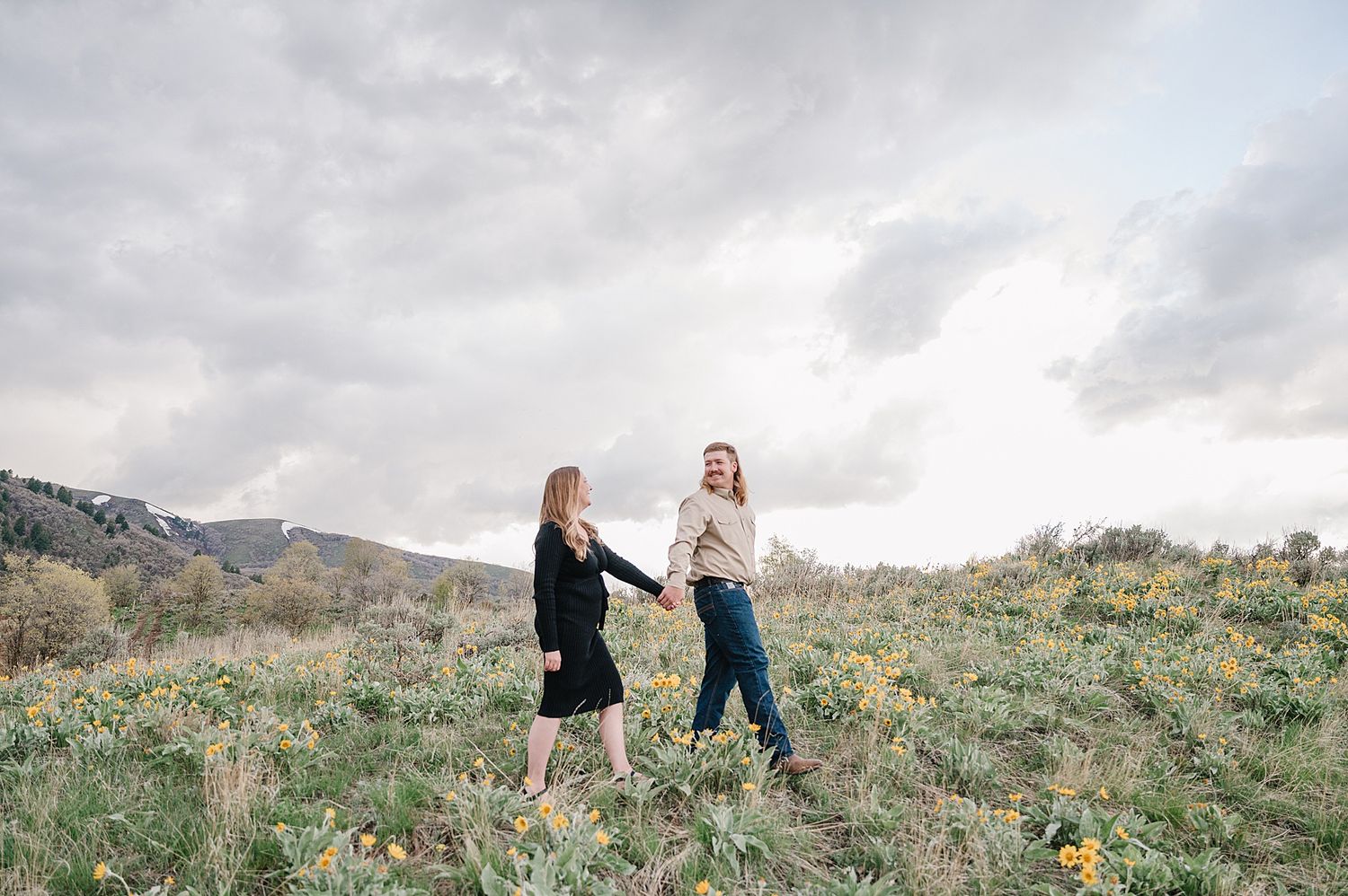 Two people holding hands while standing on a grassy hillside under moody cloudy skies in outdoor nature setting.