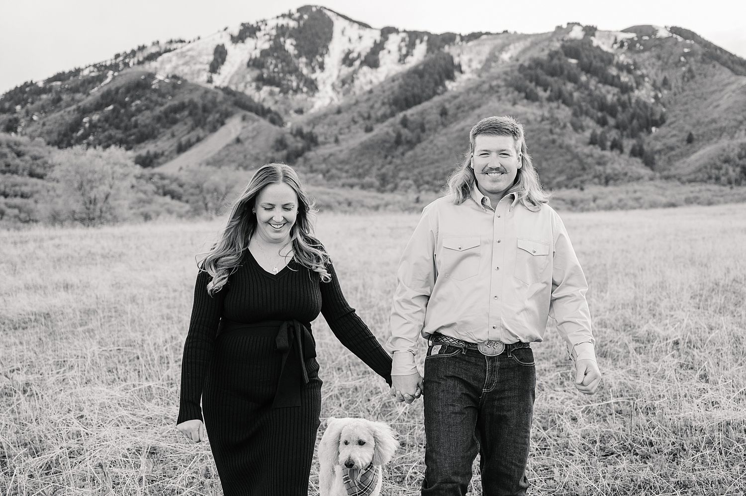 A couple walks together with their golden retriever in a scenic mountain meadow.