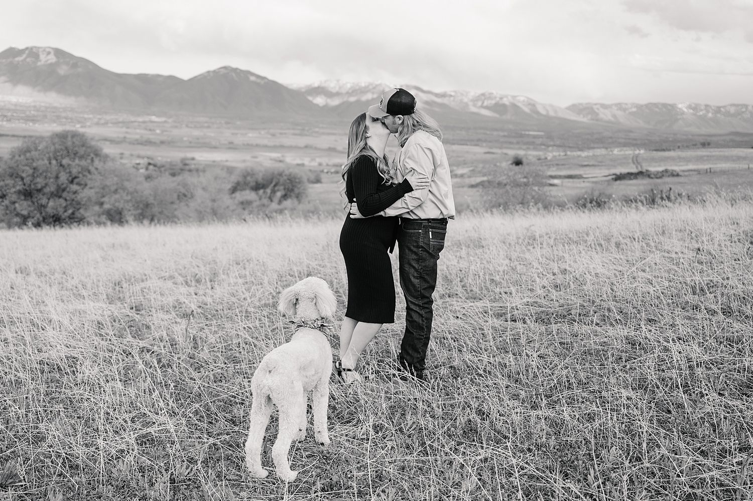Couple walking together in a grassy field with their white dog against a mountain backdrop in black and white.