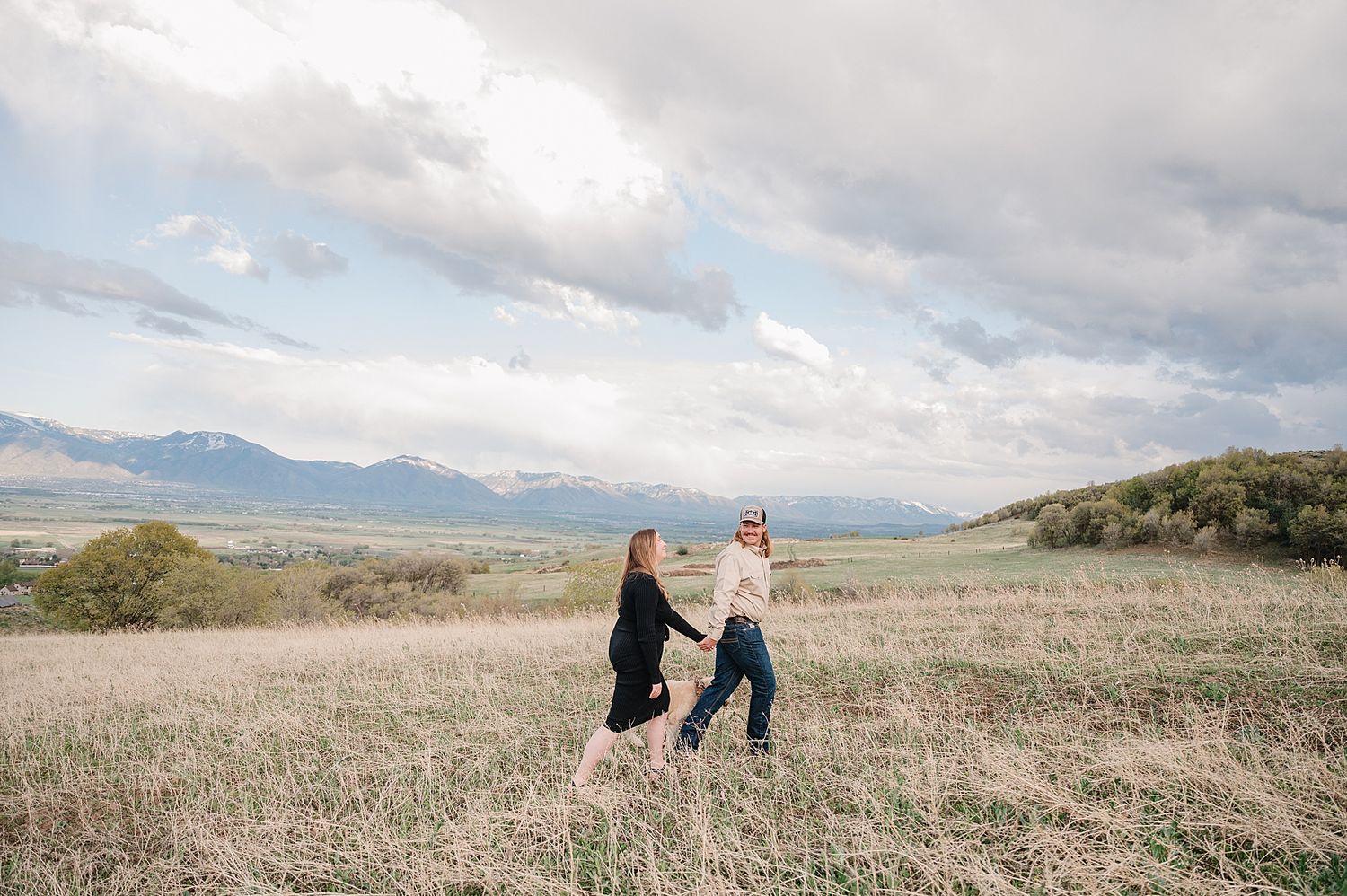 Two people dancing together in a grassy mountain meadow with dramatic cloudy skies and mountain peaks in the distance.