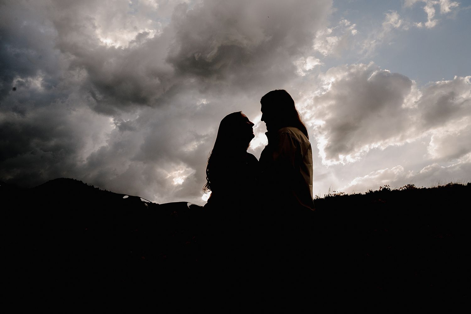 A silhouetted couple stands close together against a dramatic cloudy sky at sunset creating a romantic mood.