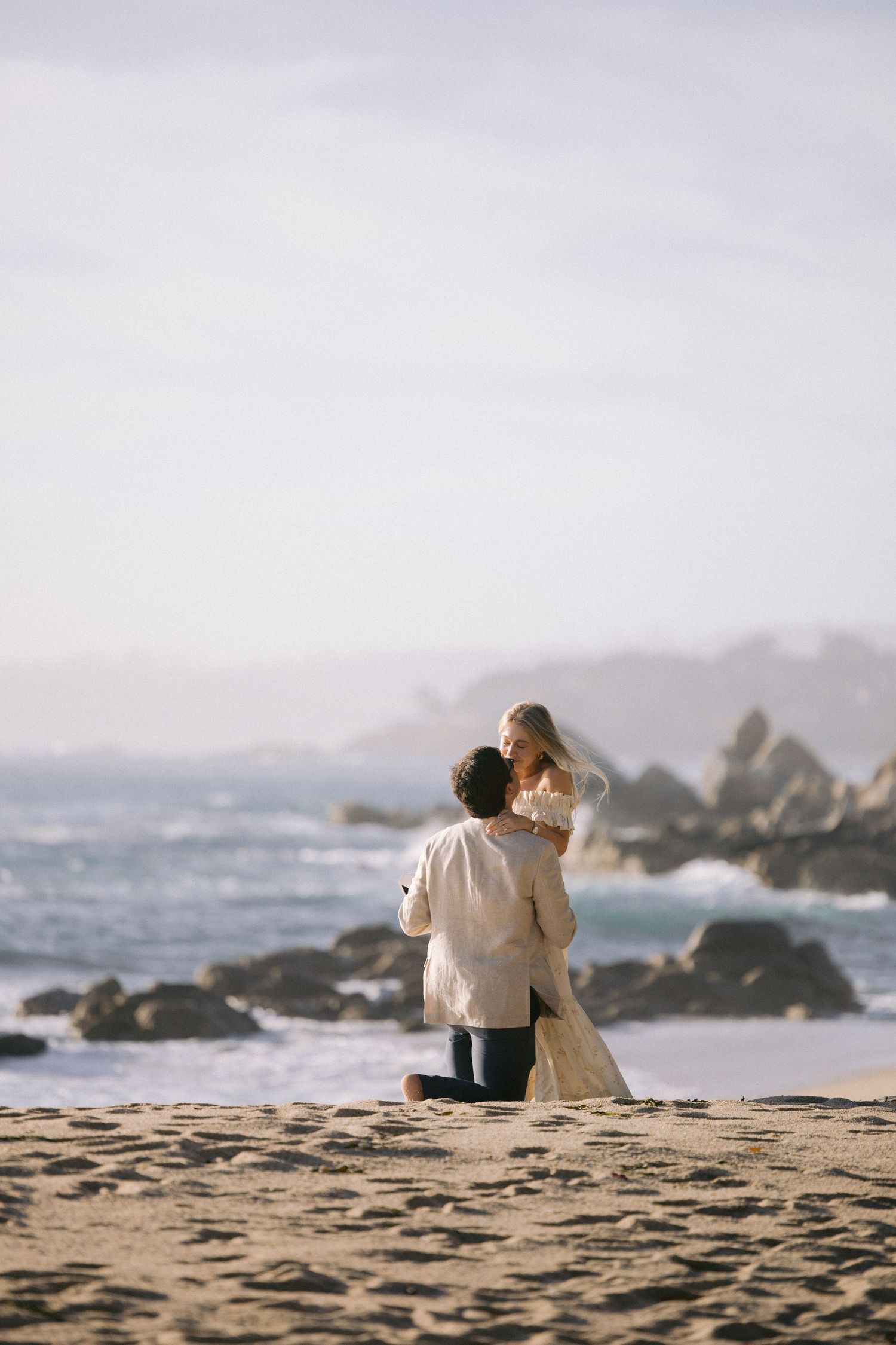 A couple stands together on a sandy beach with rocky coastal cliffs and crashing waves in the background.