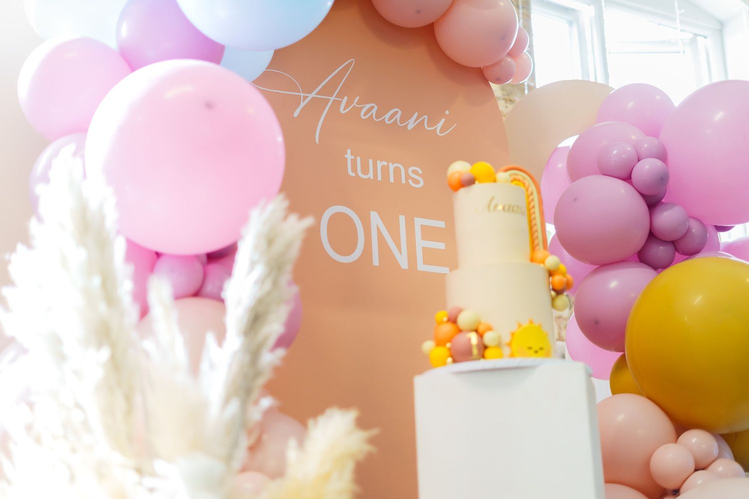 First birthday party celebration with peach backdrop, balloon garland in pink and purple tones, and white tiered cake.
