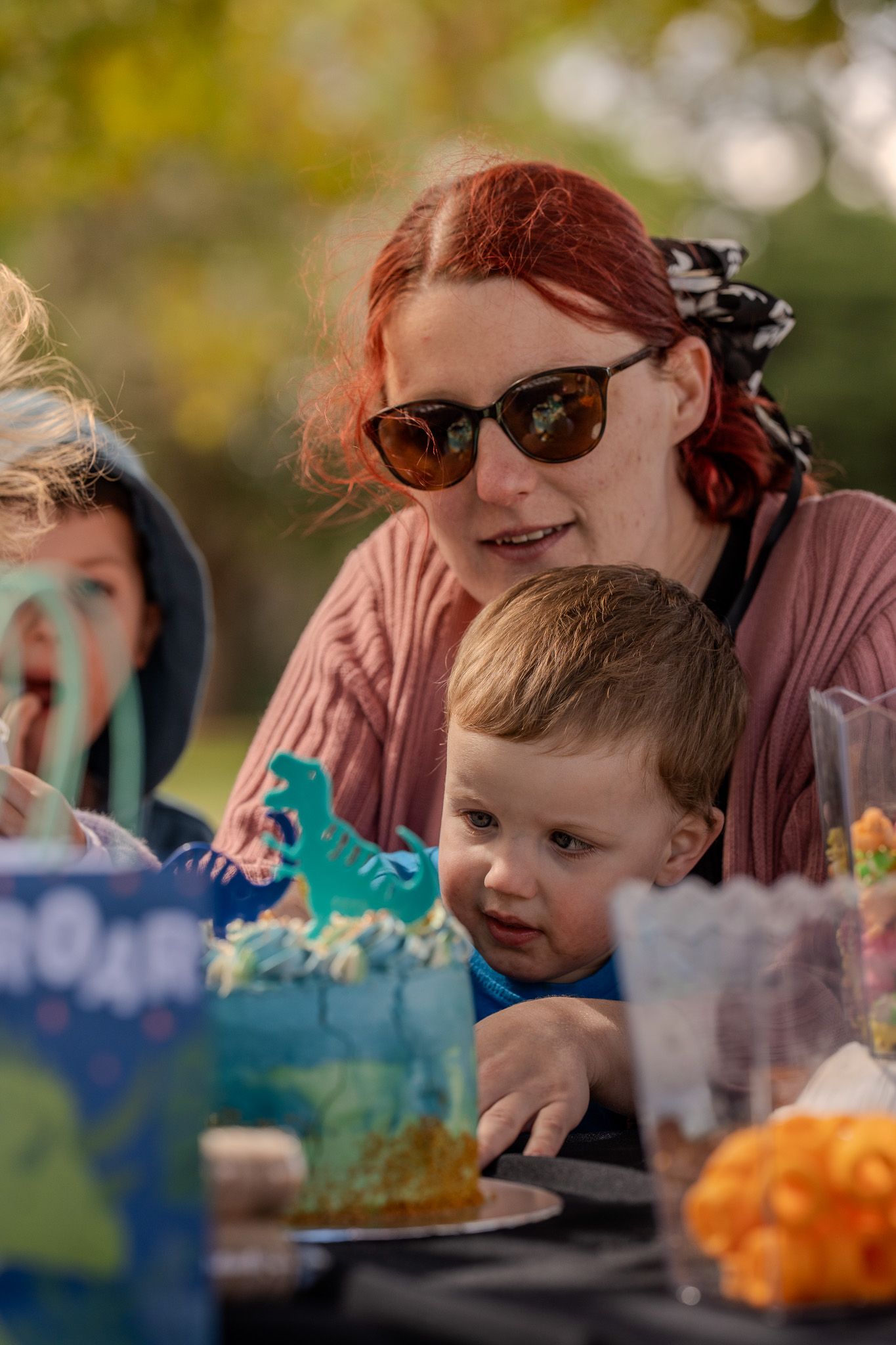 A child and adults gathered around a table at a birthday party with a dinosaur theme and colorful decorations.