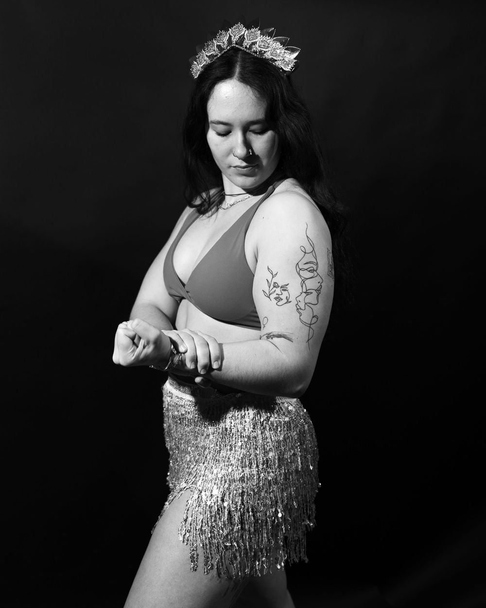 A black and white portrait of a woman flexing her bicep. She is wearing a crown and a sparkly skirt