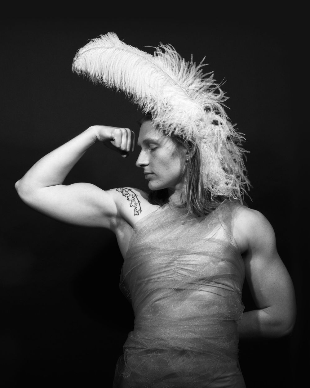 A black and white portrait of a woman flexing her bicep from the front. the woman is wearing a feather on her head