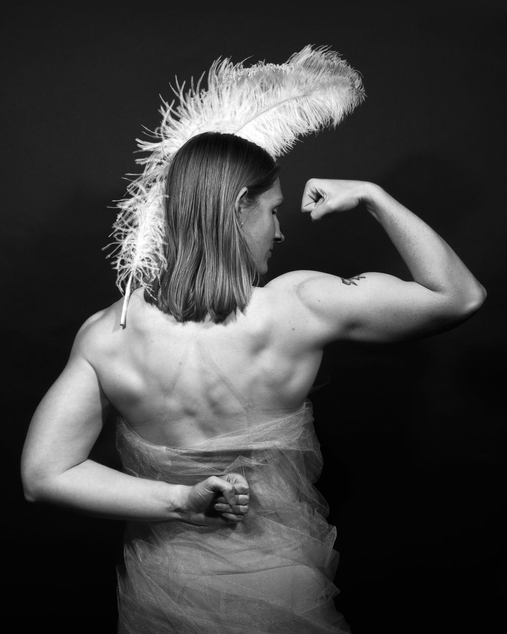 A black and white portrait of a woman flexing her bicep from the back. the woman is wearing a feather on her head