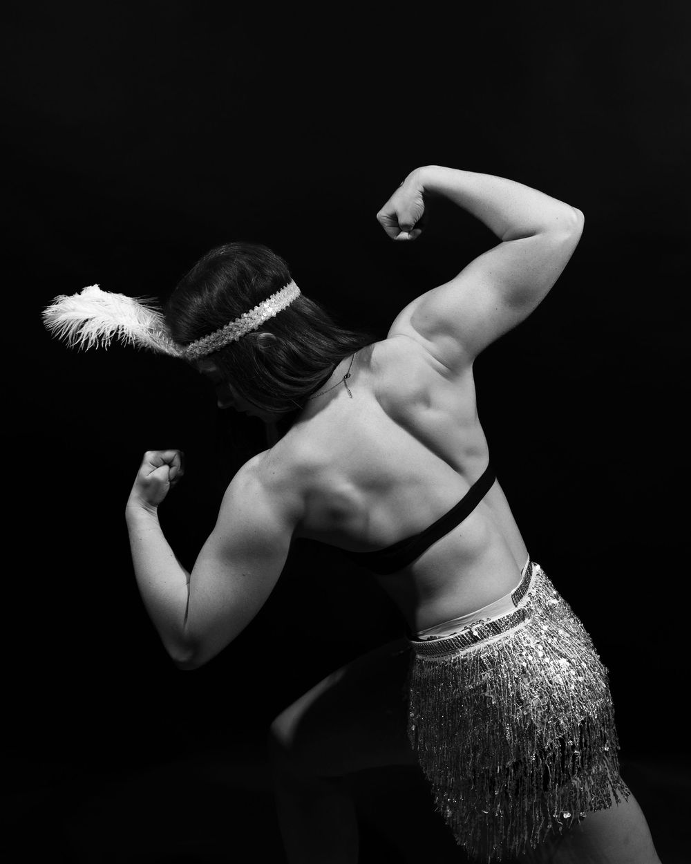 A black and white portrait of a woman flexing her back muscles. She is wearing a feather on her head and a sparkly skirt.