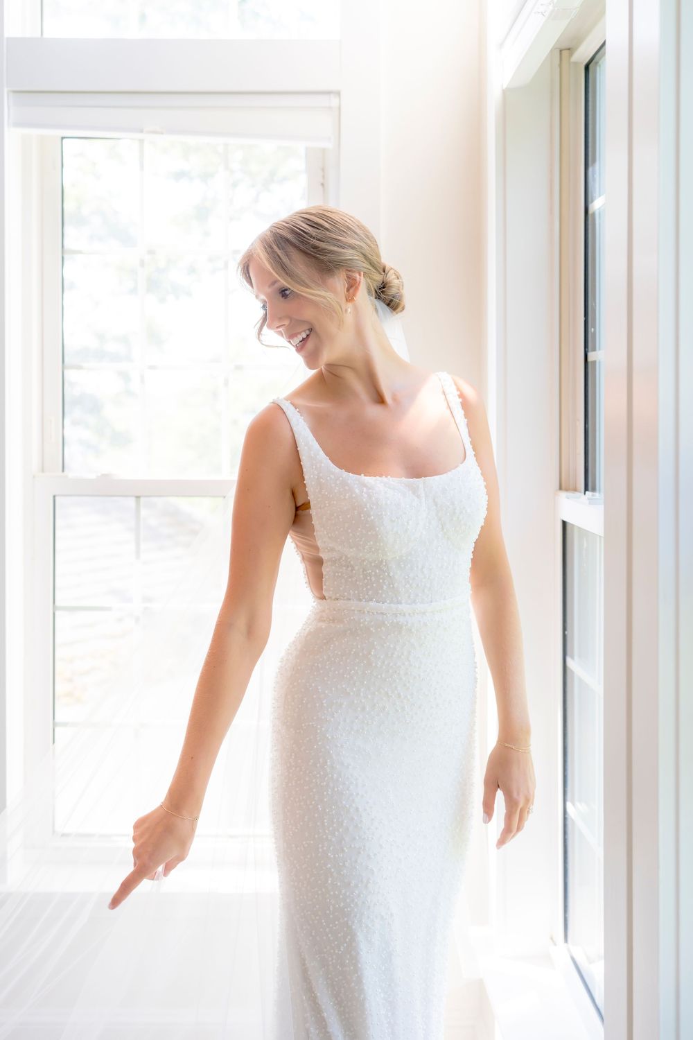 A person wearing a fitted white dress stands in a bright, sunlit room with large windows.