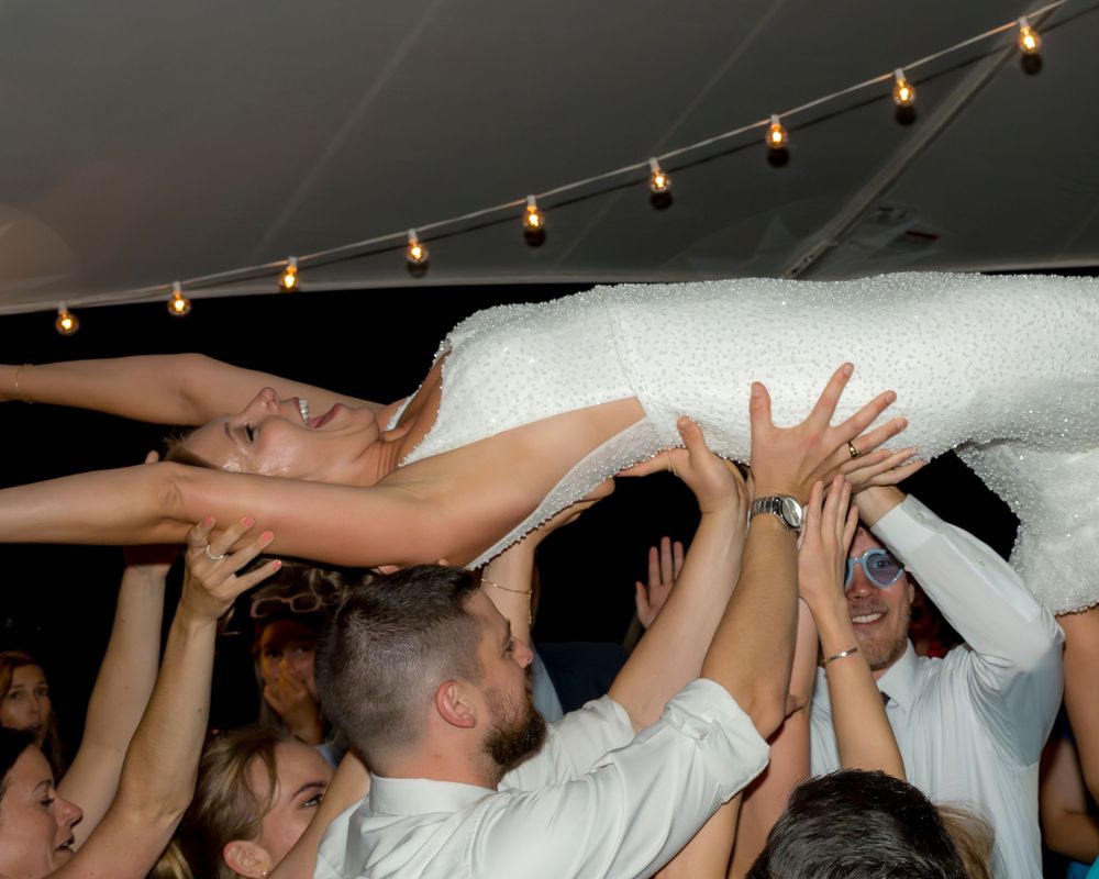 A crowd lifts someone wrapped in white fabric during a celebration with string lights overhead.