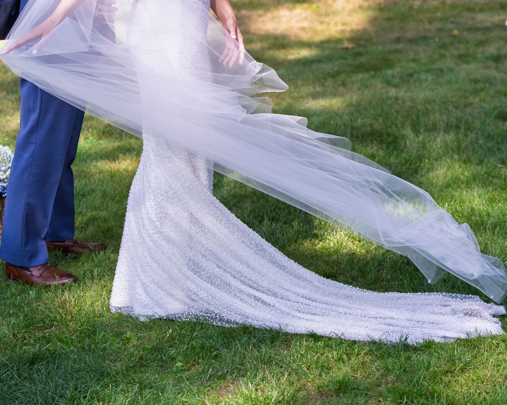 A flowing white wedding dress train trails across green grass on a sunny day.