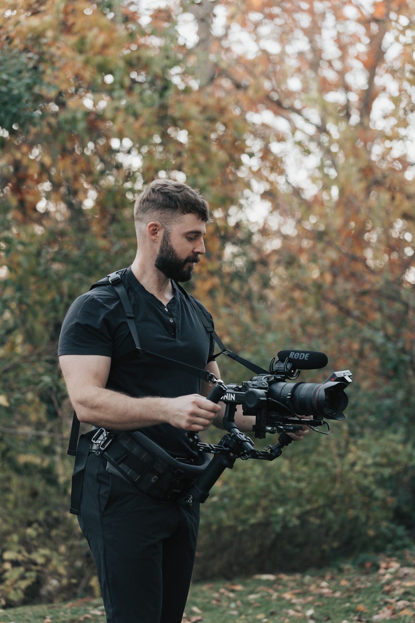 Professional photographer in black attire aims camera with telephoto lens in outdoor autumn setting.
