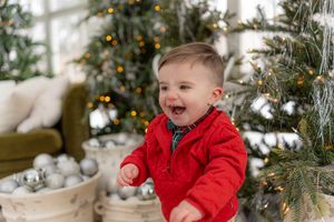 A child in a red coat plays near a decorated Christmas tree with ornaments and lights.