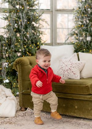 A toddler in a red sweater and khakis stands excitedly by a green velvet couch and Christmas tree.