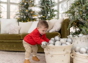 A toddler in red reaches into a white container filled with silver ornaments near a Christmas tree.