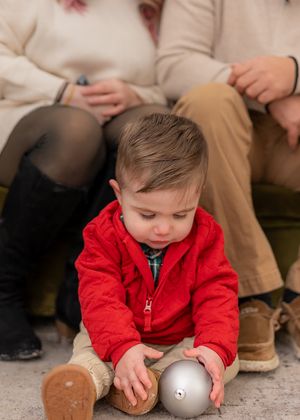 A small child in a red jacket plays with silver Christmas ornaments on the floor.