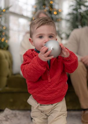 A toddler in a red sweater holds up a silver ornament ball during holiday decorating.