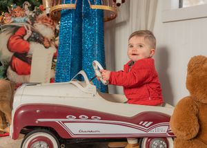 A child in red plays in a vintage-style toy pedal car near Christmas decorations.
