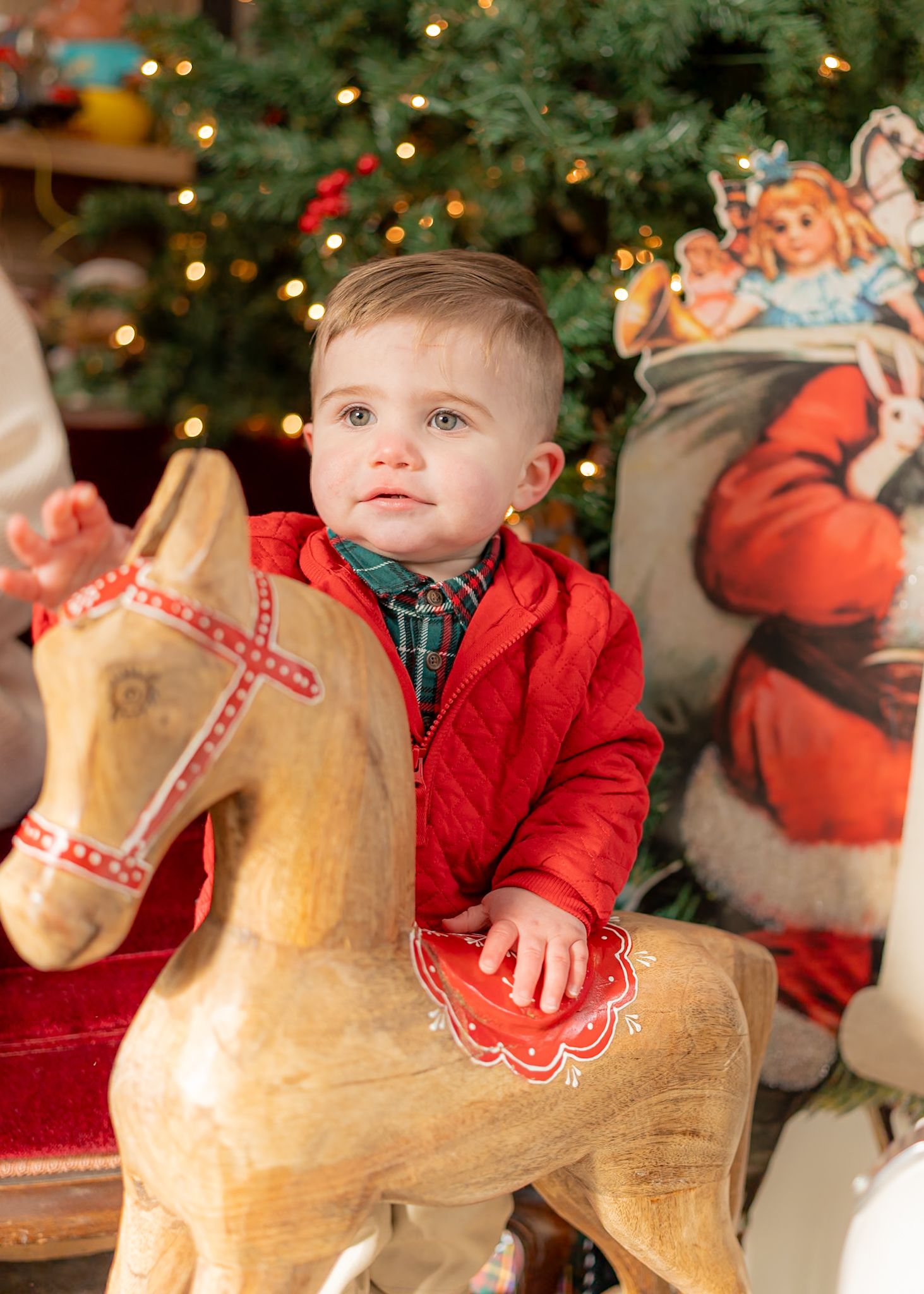 A child in a red jacket sits on a decorative wooden rocking horse near a Christmas tree.