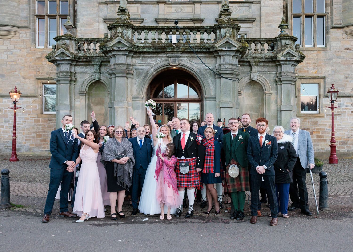 Wedding party poses for formal group photos outside historic stone building with ornate architecture.