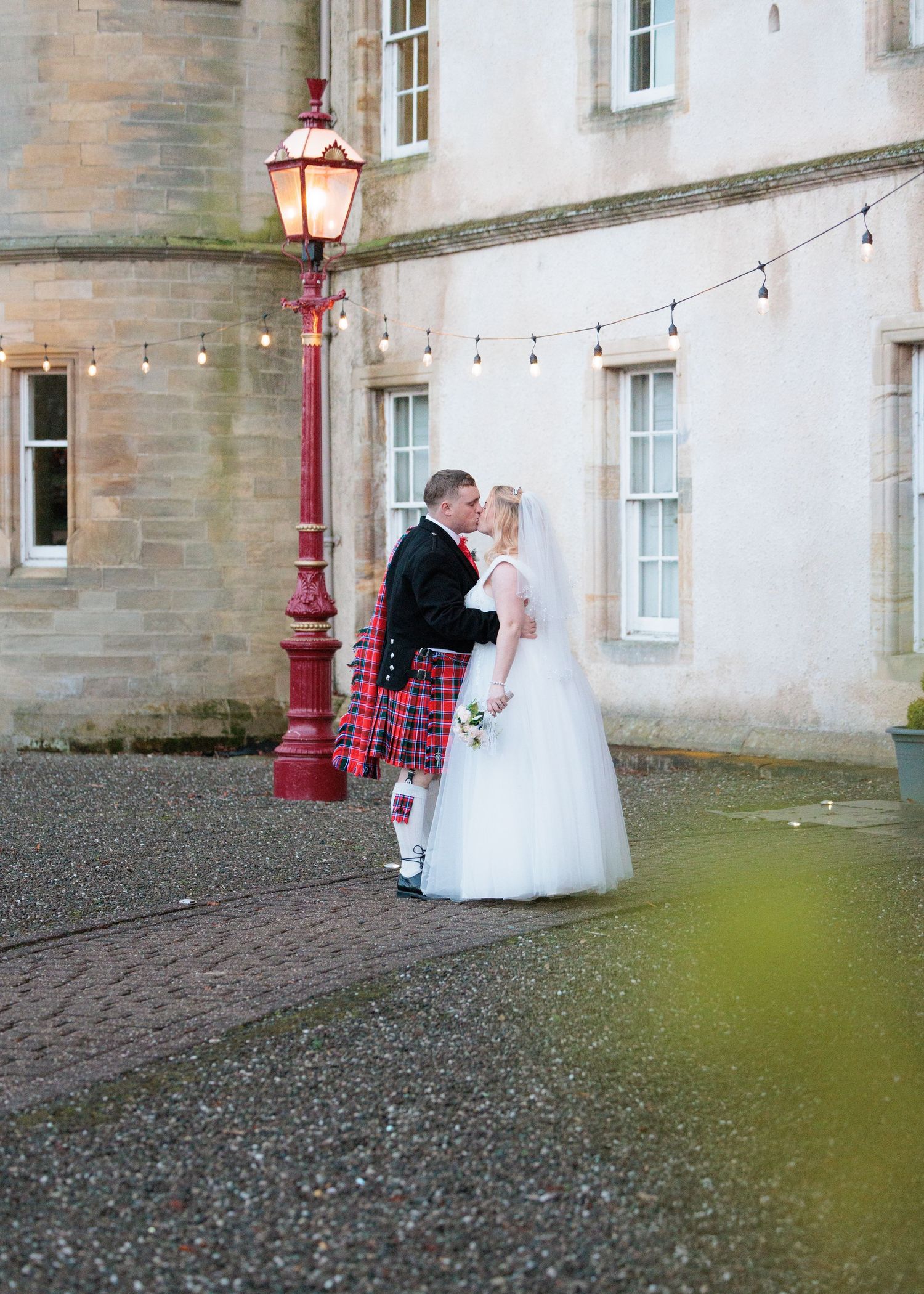 Scenic outdoor wedding photos being taken at a stone building with string lights.