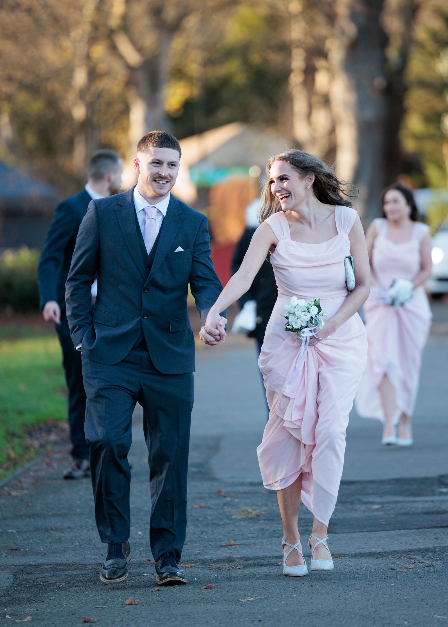 Wedding party members in formal attire run playfully down a tree-lined path during an outdoor photo session.