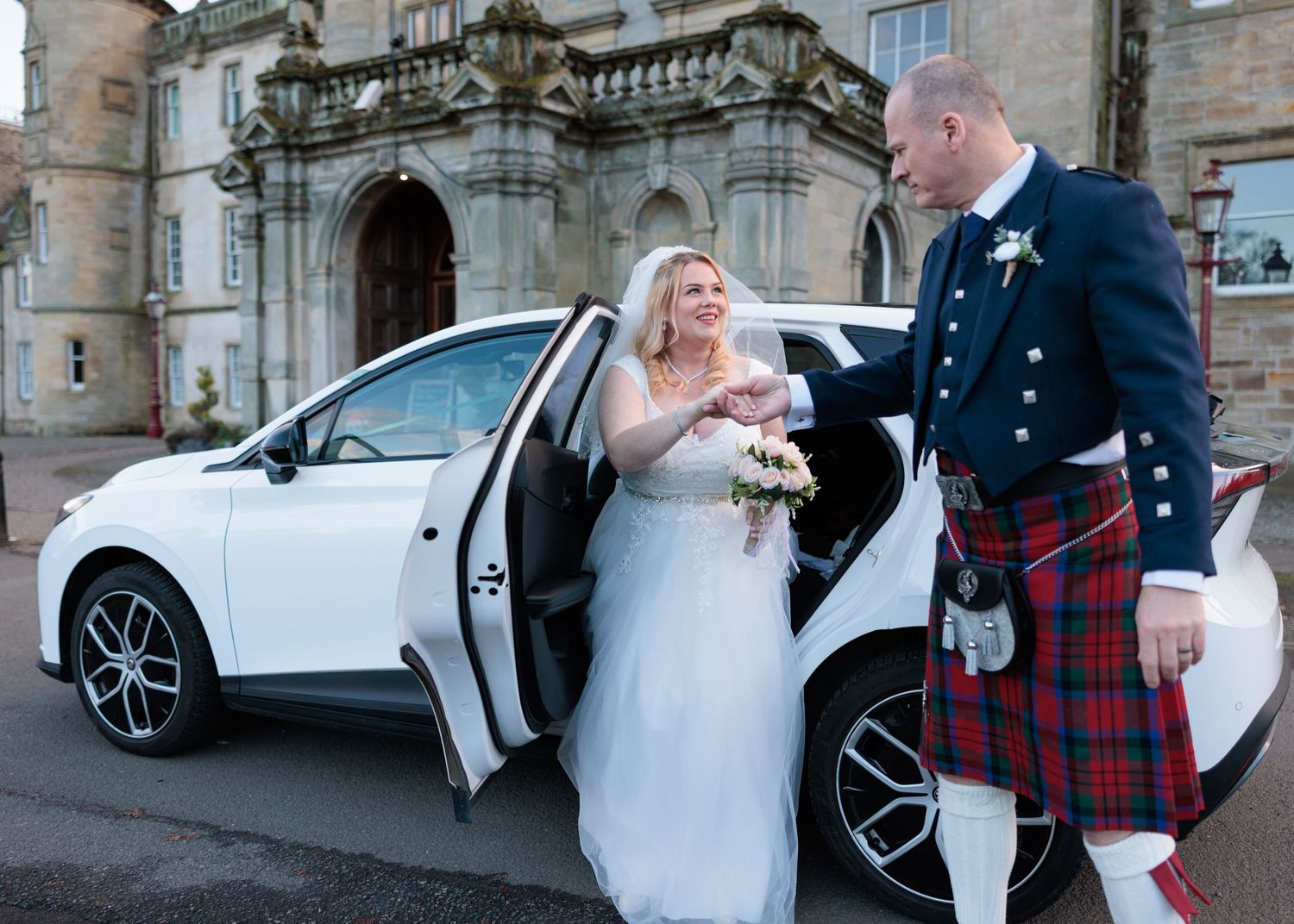 A wedding couple exits a white car outside a historic stone building, with the groom wearing traditional Scottish kilt attire.