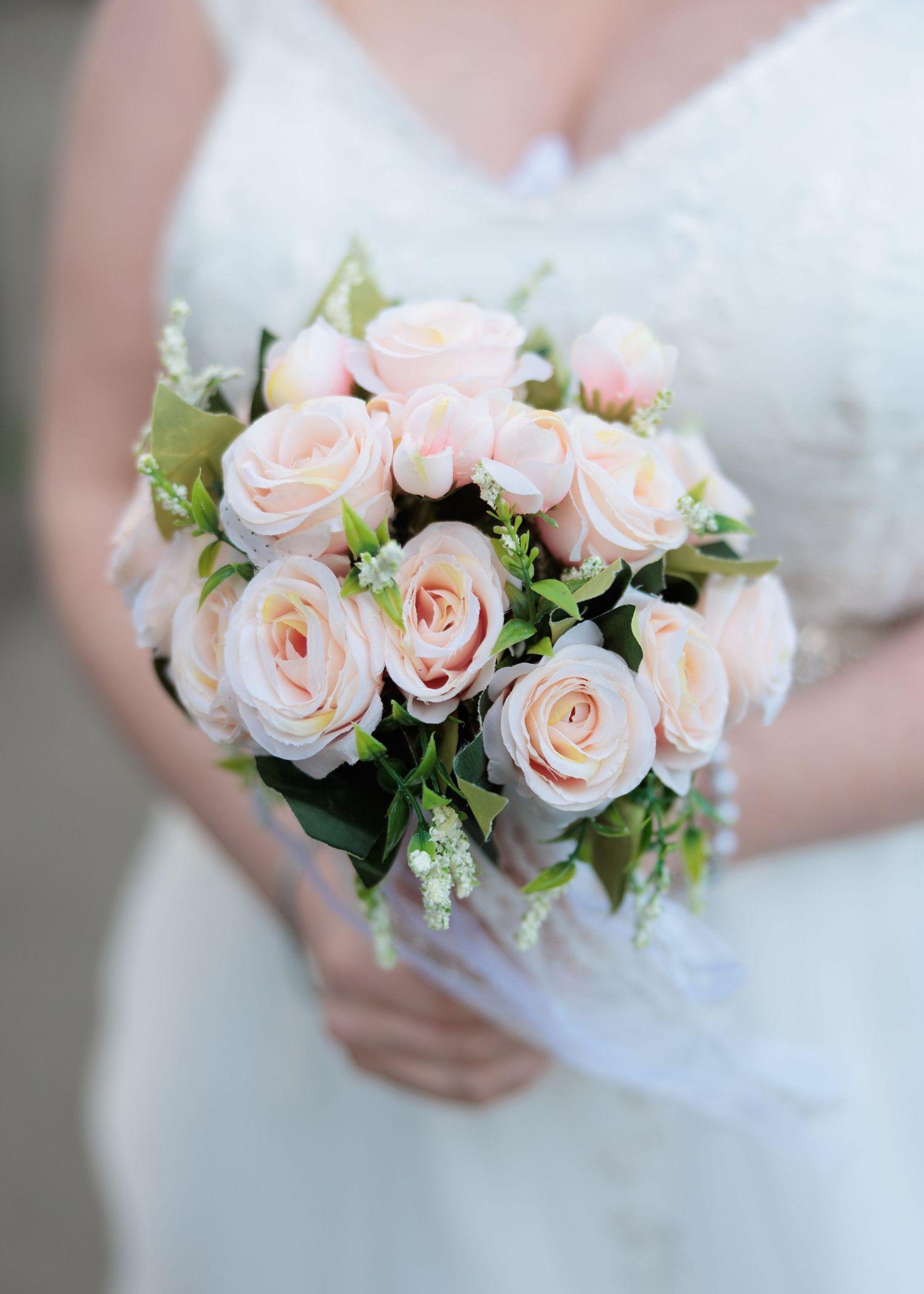 A delicate bridal bouquet featuring blush pink roses and greenery held against a white wedding dress.