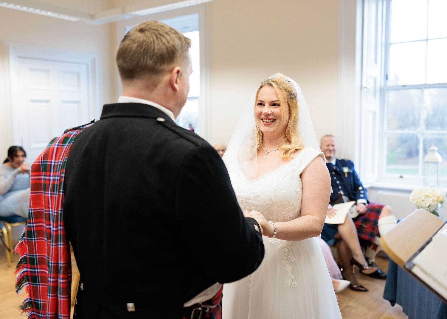 Wedding party members laugh and socialize in a bright white room with large windows.