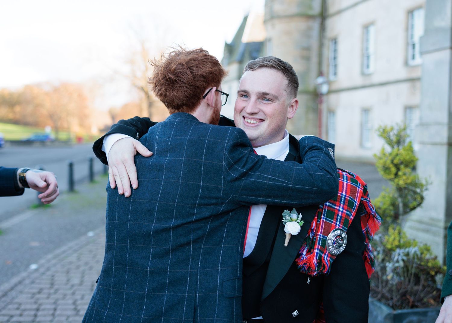 Groomsmen share joyful moments and embraces outside a stone building during a wedding celebration.