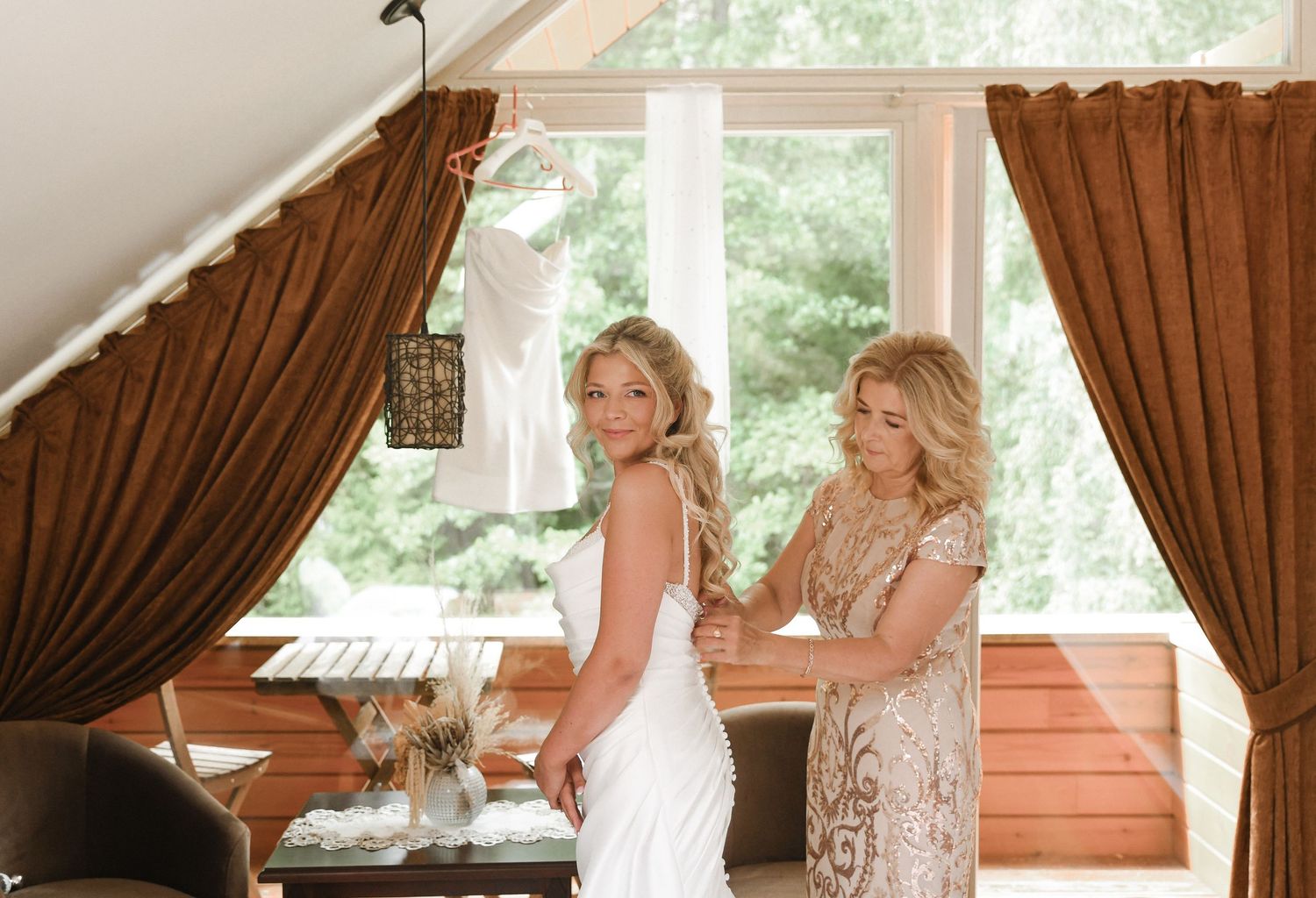A bride and her companion share a special moment in a room with brown curtains and natural lighting.
