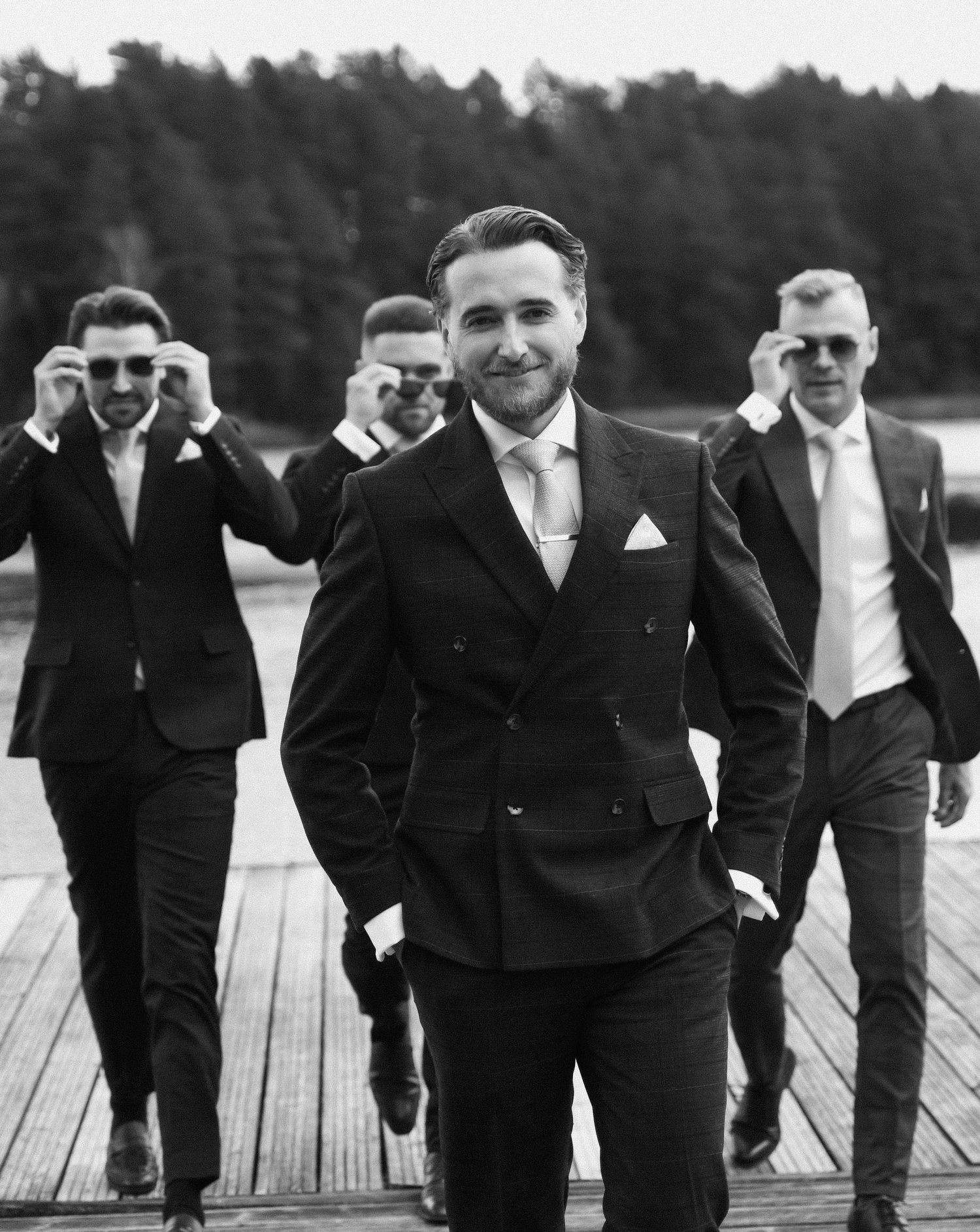 Groomsmen in black tuxedos pose together on a wooden dock in a creative panoramic wedding photo.
