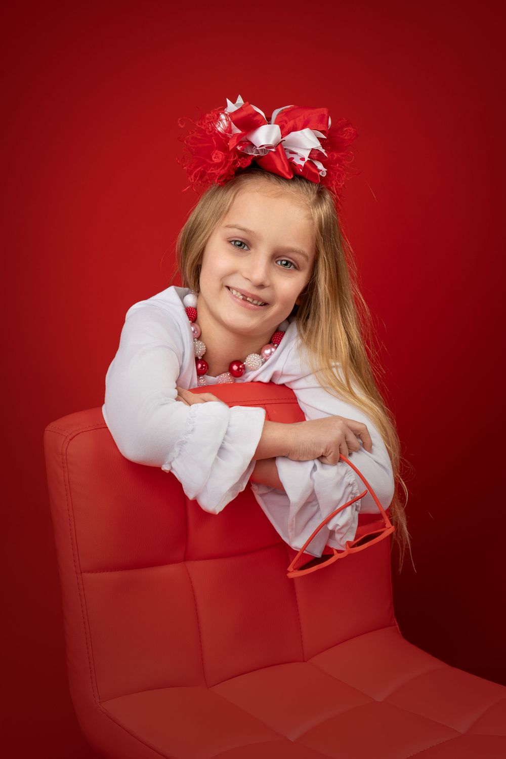A child in a white dress rests on a red couch against a vibrant red background, creating a festive holiday portrait.