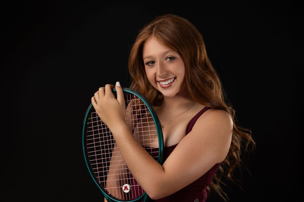 A tennis player smiles while holding a racquet against a dark background.