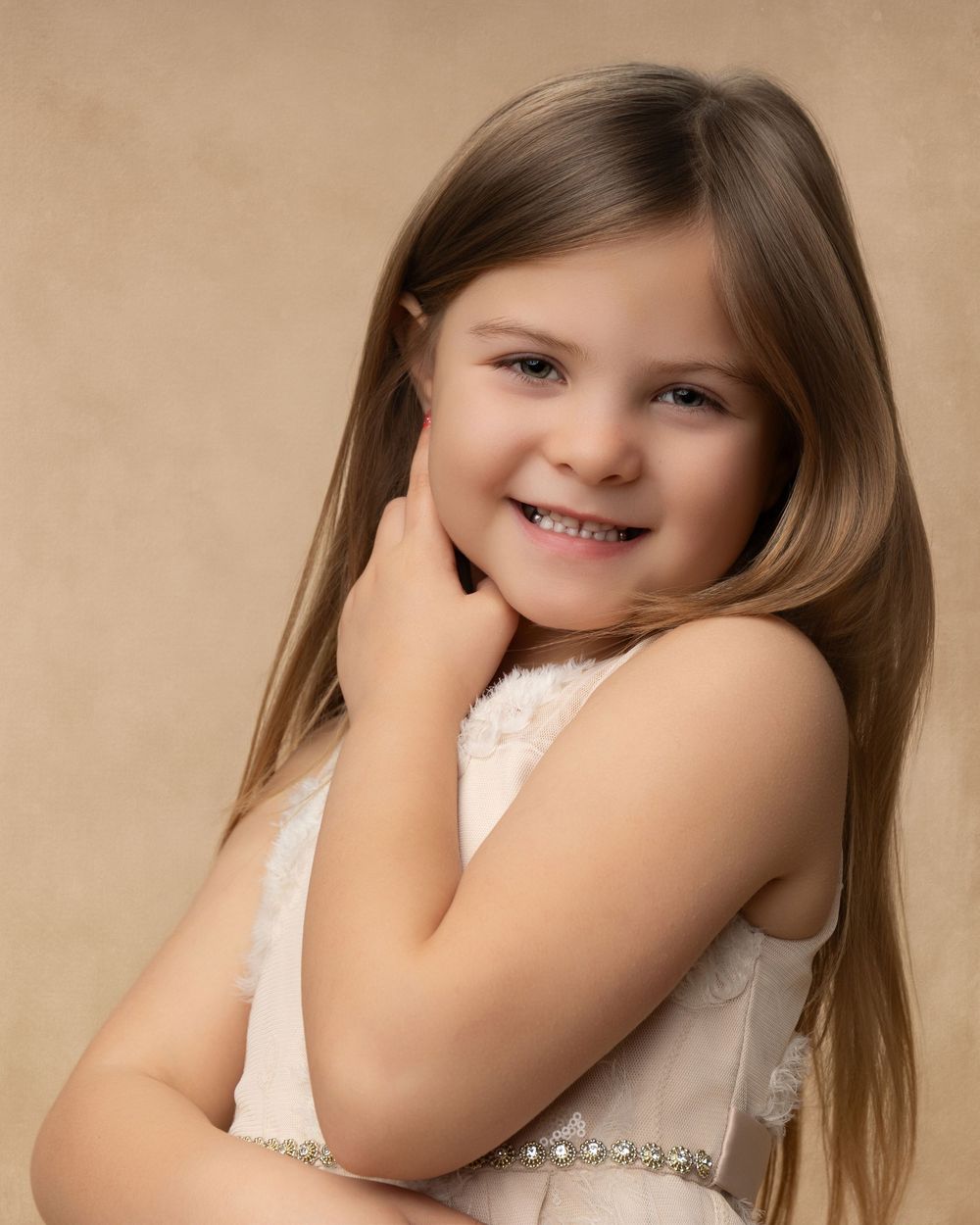 A young child in a white lace dress poses with long brown hair and a bright smile against a beige background.