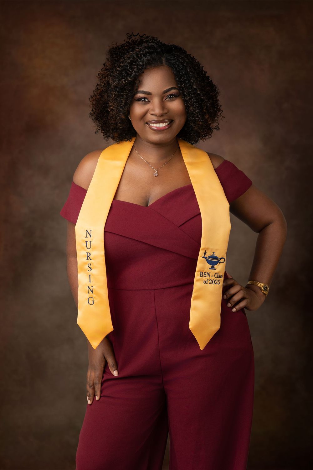Graduation portrait in burgundy jumpsuit with gold honors stole against brown backdrop.