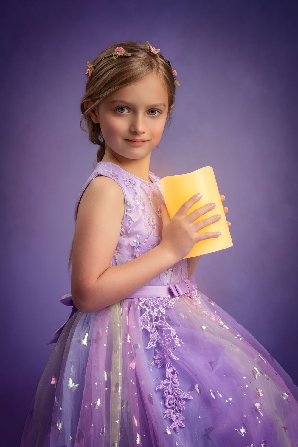 Young girl in purple princess gown holding glowing yellow book, with floral hair accessories, on lavender background.
