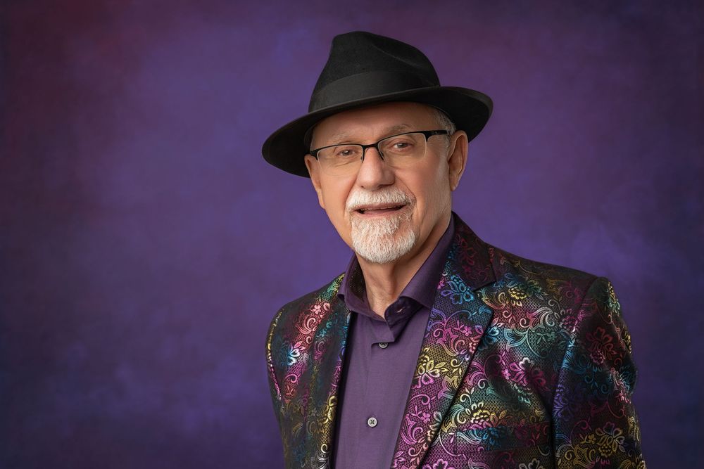 Elderly man wearing colorful floral blazer and black fedora hat, smiling against purple studio background.