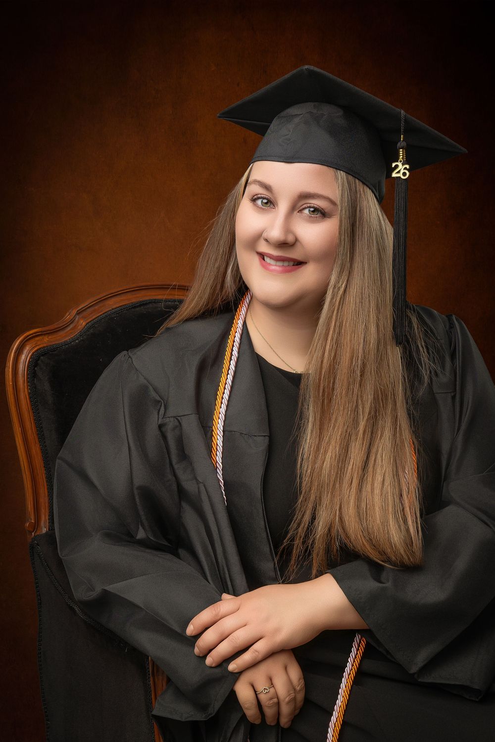 Smiling graduate in black cap and gown with gold cords seated in ornate chair for formal portrait photo.