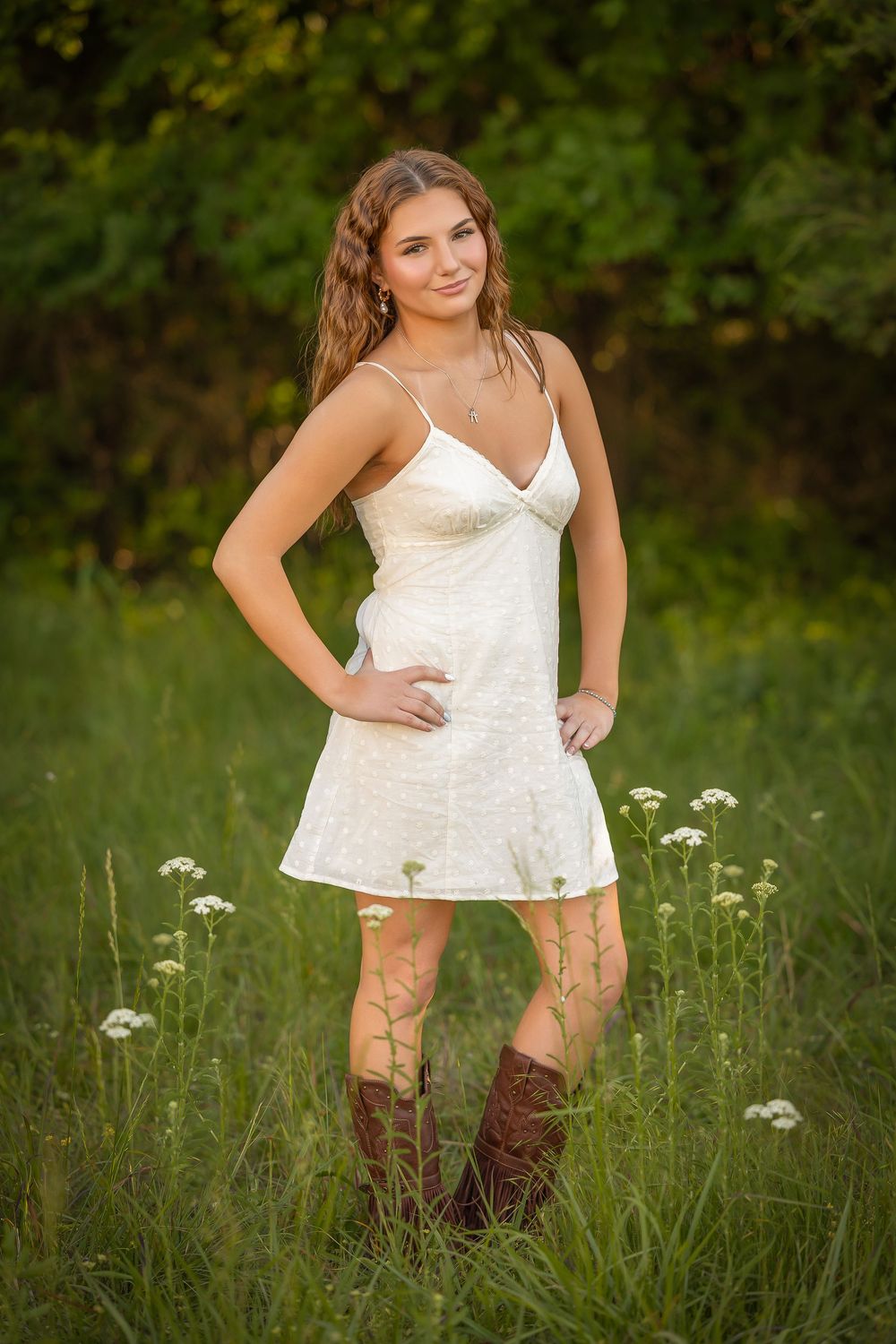 Young woman in white dress and brown cowboy boots standing in green field with wildflowers.