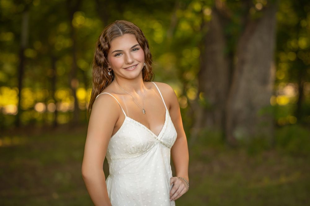 Young woman in white lace dress smiling outdoors in a sunlit green forest setting.