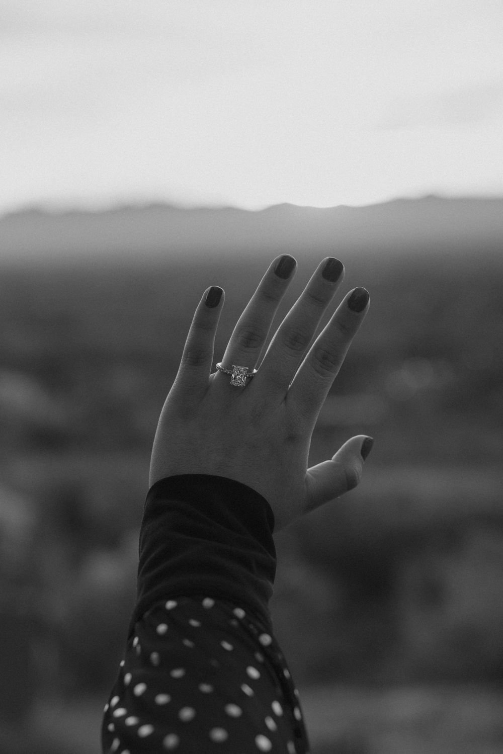 A hand wearing a polka dot sleeve raises against a misty mountain landscape, displaying an engagement ring in black and white.