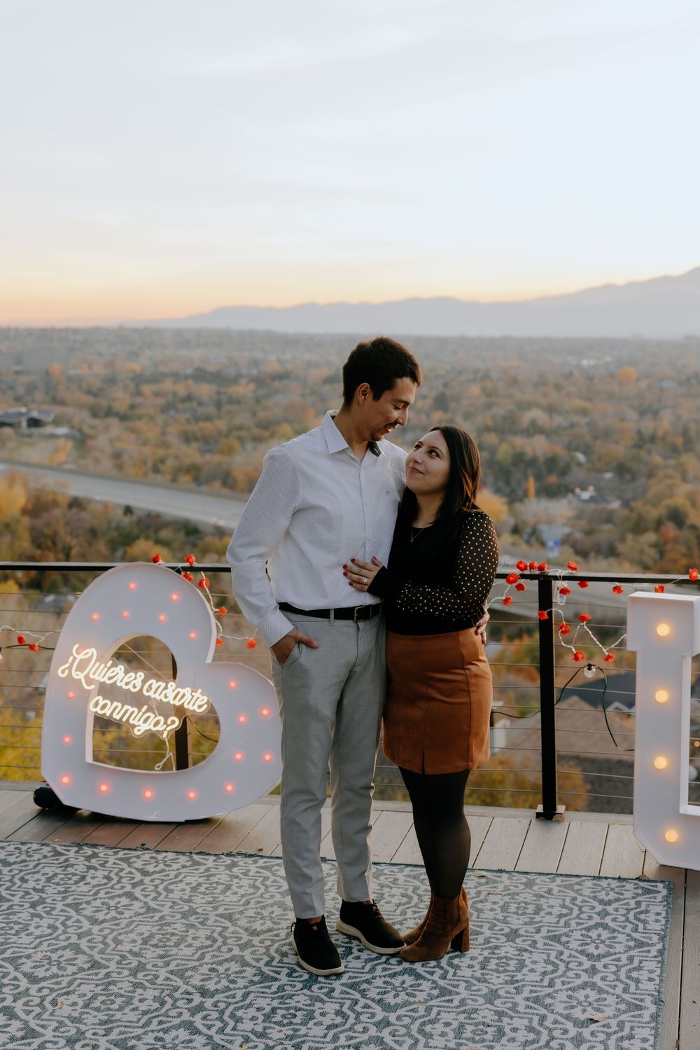 A romantic proposal setup on a rooftop terrace with glowing letters and string lights overlooking a scenic autumn landscape.